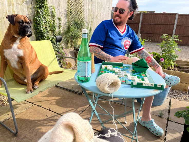 A photo of an adult boxer dog sitting outside on a green patio sofa. Next to him is a white male with sunglasses and wearing blue crocs. He sits across the table from the photographer who is knitting. On the table in the middle, a game of scrabble is in progress.