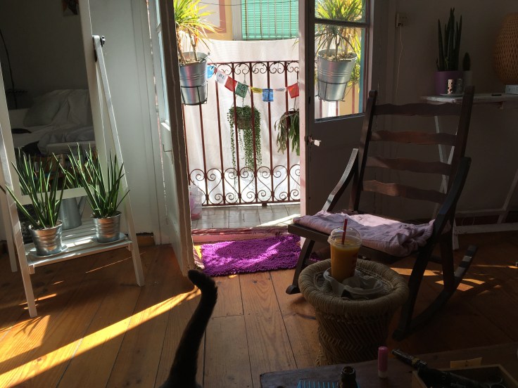 Shot of a Spanish living room. Balcony doors are open leading to a small balcony with some hanging plants and balcony planters and some Tibentan prayer flags add some colour. The balcony overlooks a narrow street. Foreground is a rocking chair and an iced drink