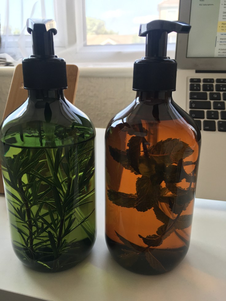 Two oil bottles side by side. One bottle is green and has a sprig of rosemary inside, one bottle is brown and has some mint sprigs inside