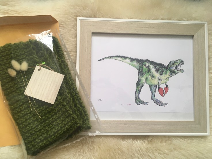 Left: a green knitted cowl in clear plastic and tree bunny tail flower stems ad gift card. Right: A4 Framed artwork of a green dinosaur roaring and clutching a broken heart.